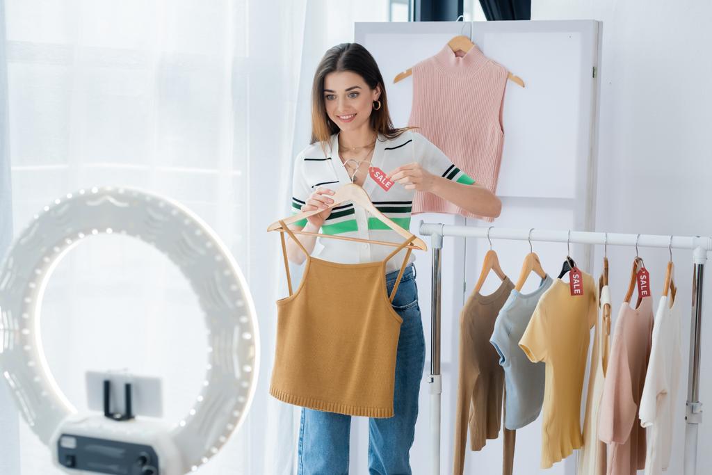 fröhliche Mode-Bloggerin hält Tag mit Verkauf Schriftzug in der Nähe von Kleidung und Handyhalter - Foto, Bild