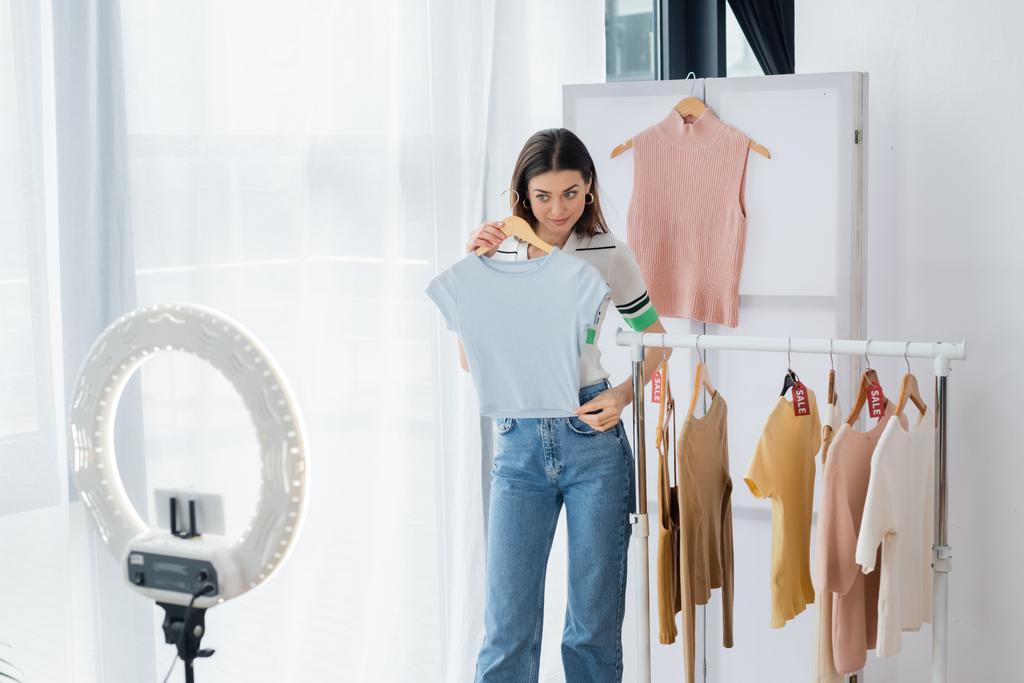 jolie blogueur de mode montrant t-shirt près du porte-téléphone avec lampe circulaire - Photo, image