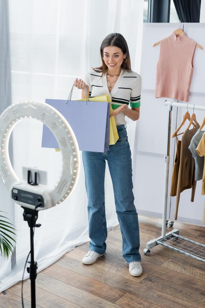 Lächelnder Modeblogger zeigt auf Einkaufstüten in der Nähe von Handyhalterungen mit Ringlampe - Foto, Bild