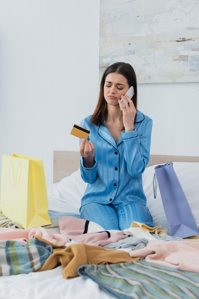 服や買い物袋の近くにクレジットカードを持っている間に携帯電話で話す絹のパジャマの悲しい女性 - 写真・画像