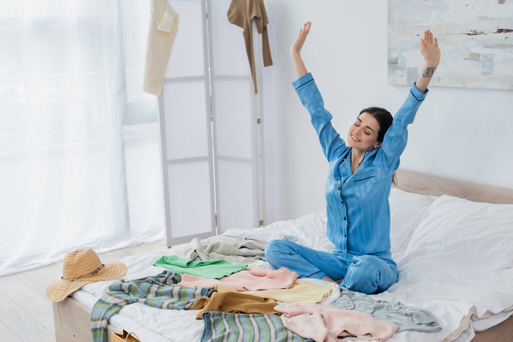 joyful woman in silk pajama stretching on bed near plenty of different clothes - Photo, Image