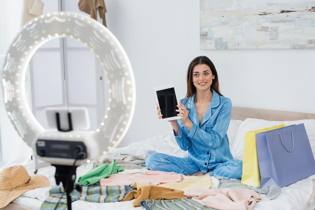 fröhliche Modebloggerin im trendigen Pyjama, die ein digitales Tablet in der Nähe eines Handyhalters mit Ringlampe zeigt - Foto, Bild