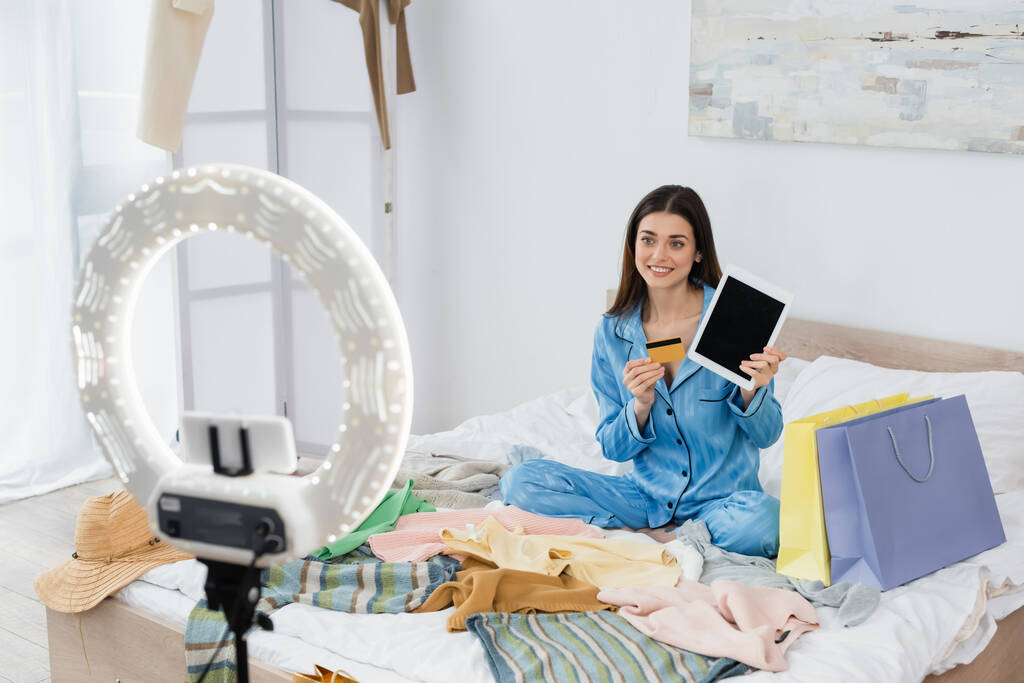 mosolygós nő elegáns pizsama digitális tabletta és hitelkártya közelében ruhák és telefon tartó gyűrű lámpa - Fotó, kép