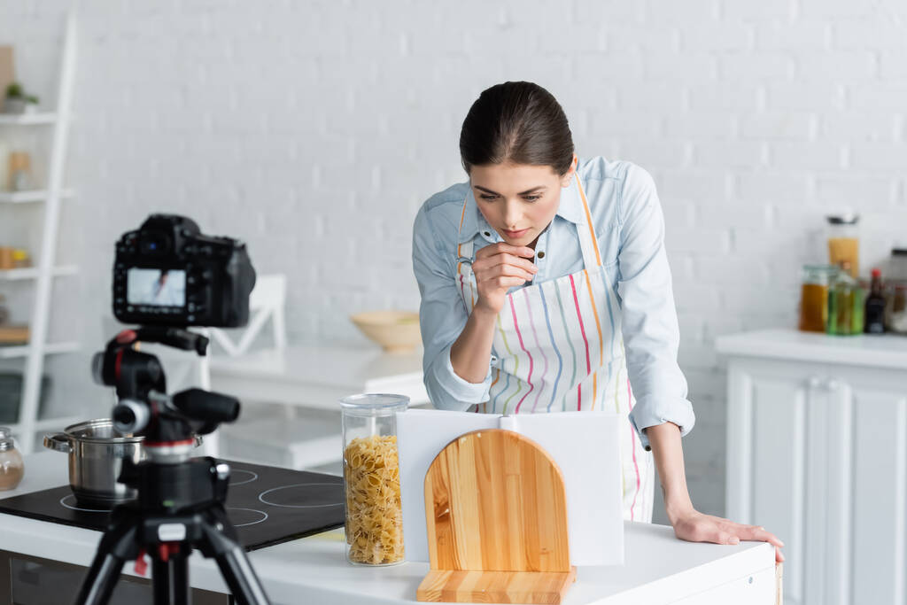 νεαρή νοικοκυρά κοιτάζοντας cookbook κοντά ζυμαρικά και θολή ψηφιακή φωτογραφική μηχανή - Φωτογραφία, εικόνα
