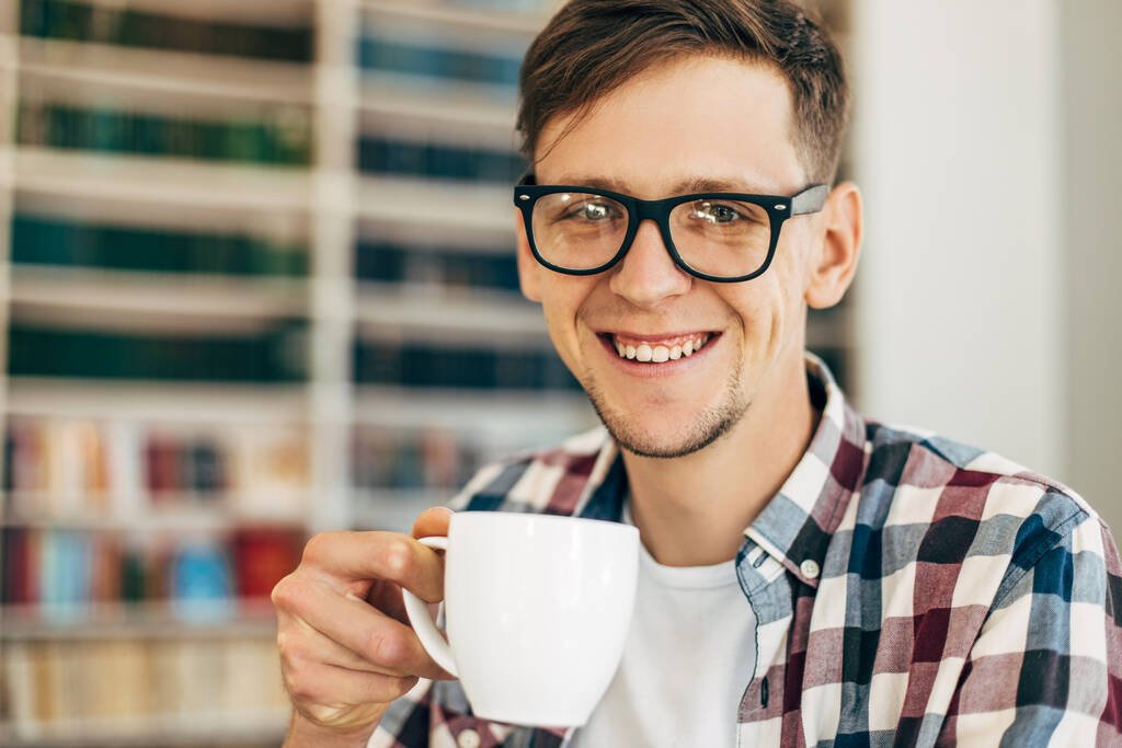 Joven sonriente con gafas disfrutando de una taza de café durante un almuerzo de descanso de trabajo en un restaurante moderno, joven inteligente relajándose en una cafetería - Foto, imagen