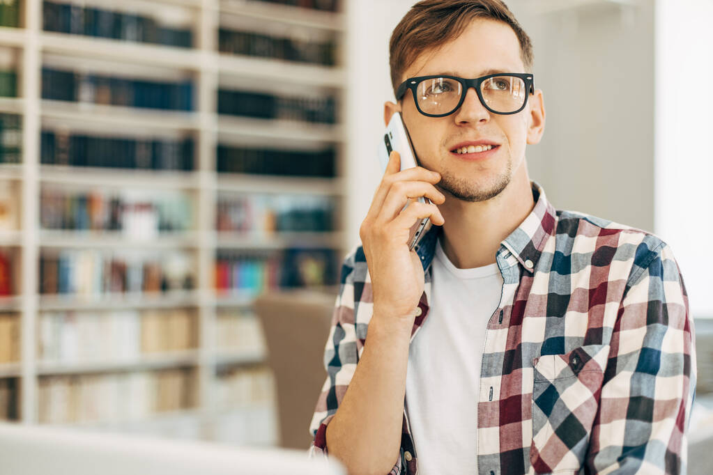 Улыбающийся бизнесмен с ноутбуком разговаривает на мобильном телефоне в офисе на фоне с библиотекой - Фото, изображение