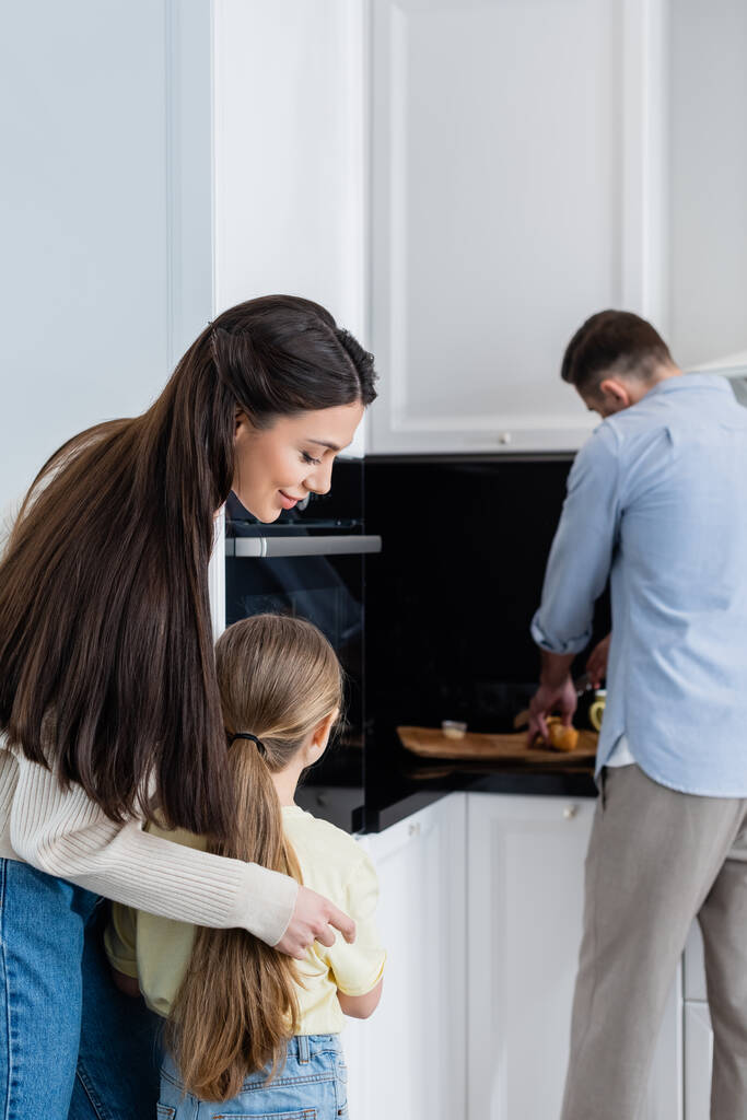 Lächelnde Frau umarmt Tochter neben verschwommenem Ehemann beim Brotschneiden in Küche - Foto, Bild