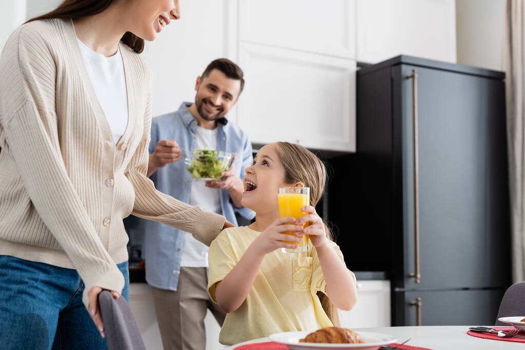 Erstauntes Mädchen mit Orangensaft neben lächelnder Mutter und verschwommenem Vater, der in der Küche Salat mixt - Foto, Bild