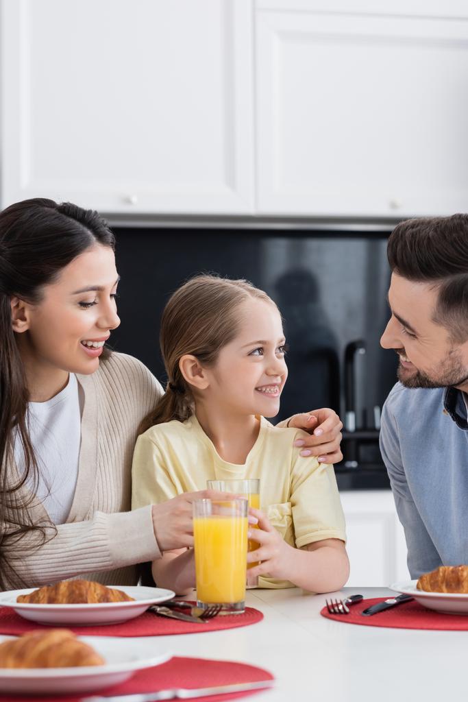 szczęśliwa kobieta obejmująca córkę przy śniadaniu i uśmiechnięty mąż w kuchni - Zdjęcie, obraz