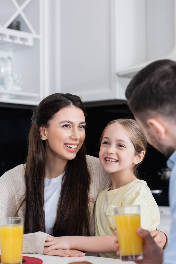 woman smiling near happy daughter and blurred husband holding glass of orange juice - Photo, Image