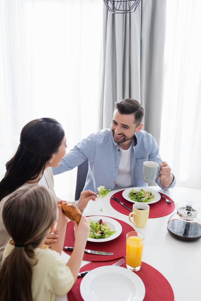 szczęśliwy człowiek uśmiechnięty w pobliżu żony i córki podczas śniadania - Zdjęcie, obraz