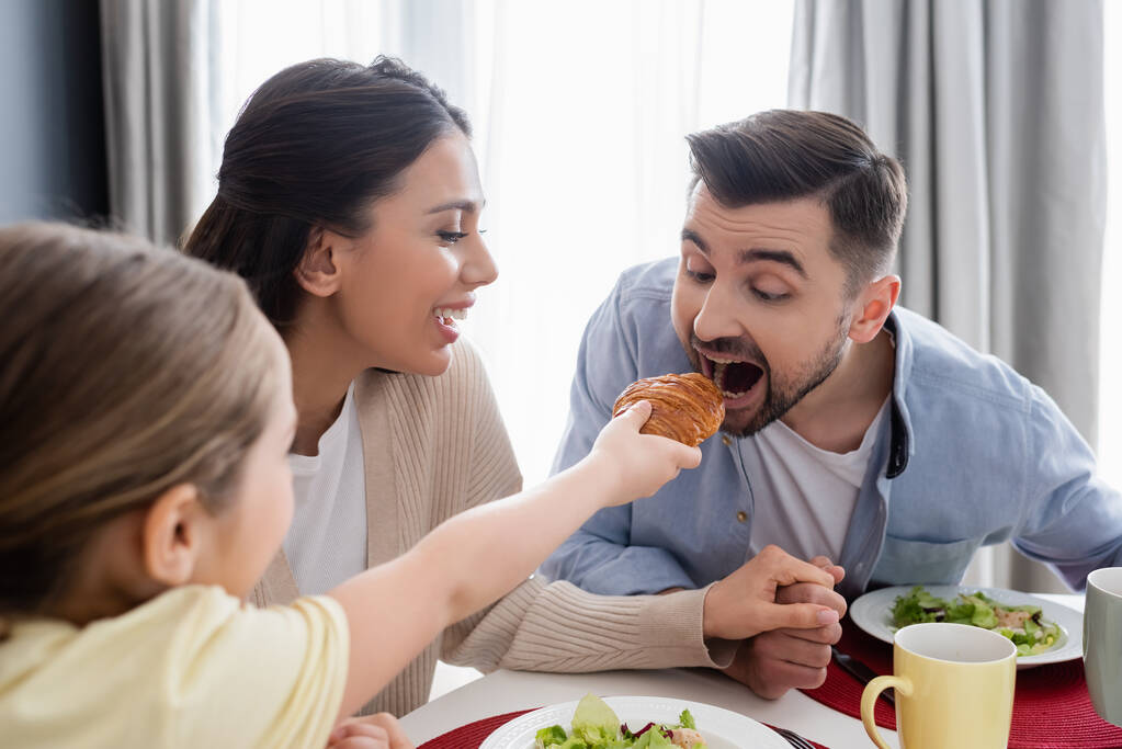 blurred child feeding dad with croissant near cheerful mother during breakfast - Photo, Image