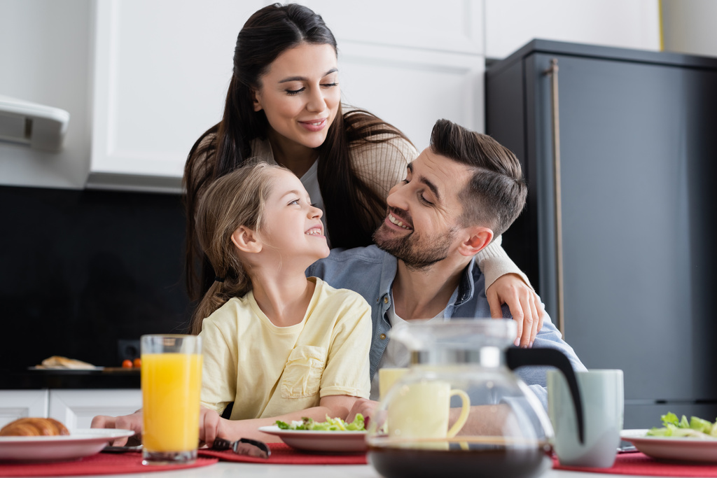 família feliz sorrindo perto do café da manhã na cozinha em primeiro plano turvo - Foto, Imagem