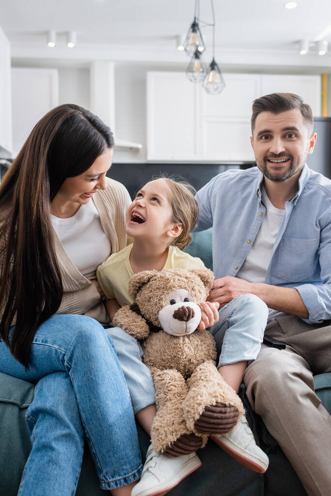ridere ragazza holding orsacchiotto mentre guardando commedia film insieme con felice genitori - Foto, immagini