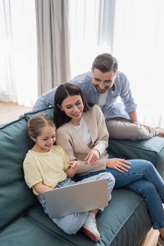 feliz hombre cerca alegre esposa e hija apuntando a la computadora portátil mientras está sentado en el sofá - Foto, Imagen
