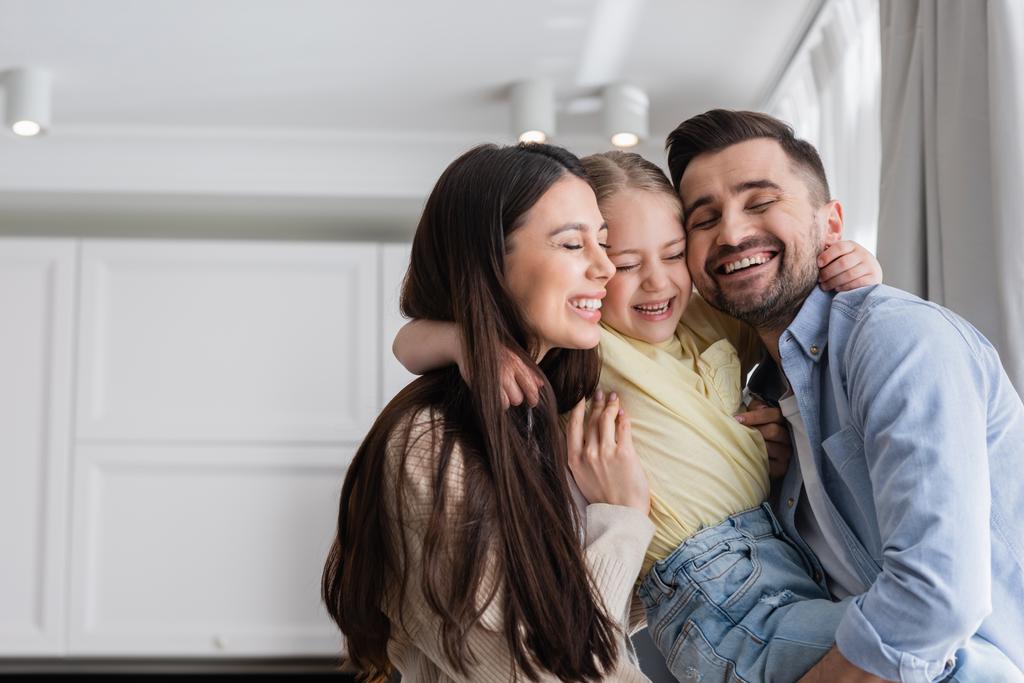 glückliche Eltern und Tochter mit geschlossenen Augen, die sich zu Hause umarmen - Foto, Bild