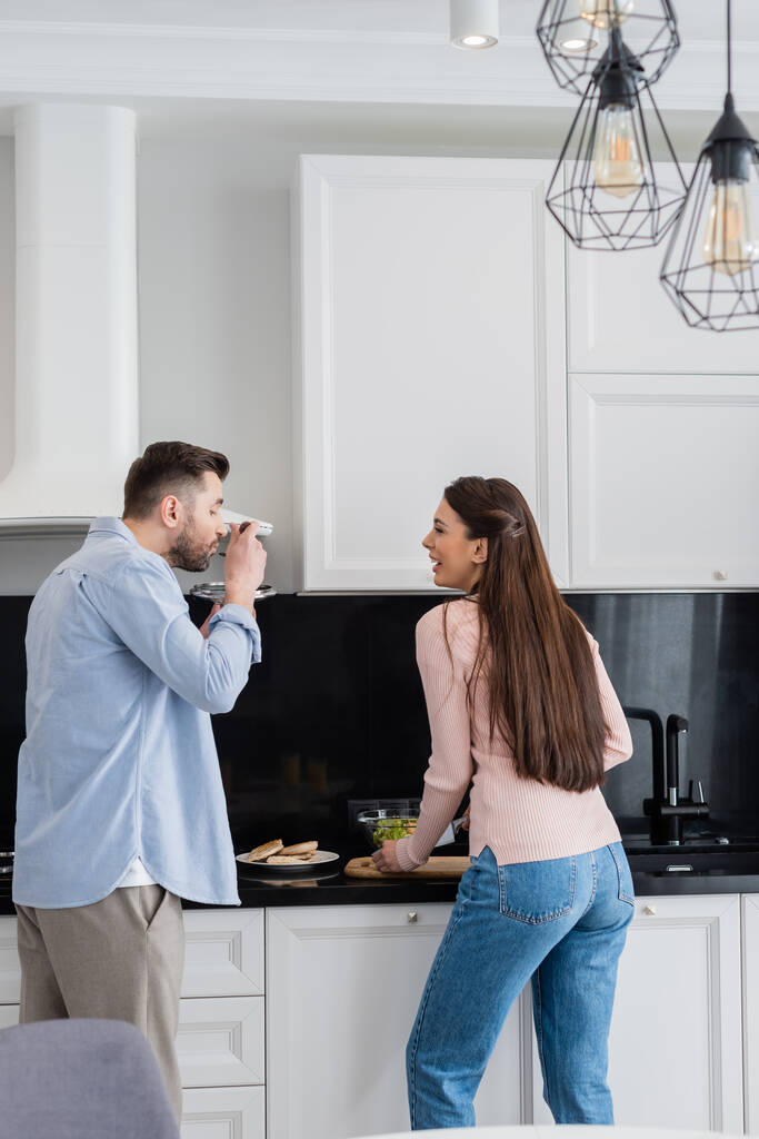 man with saucepan lid tasting meal near laughing wife in kitchen - Photo, Image