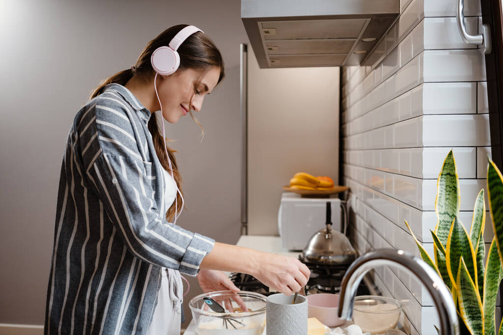 Belle femme agréable dans les écouteurs souriant et faire des crêpes pâte à la maison cuisine - Photo, image