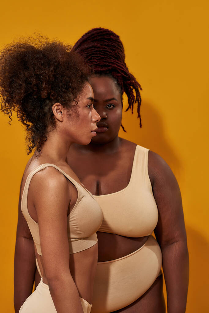 体の大きさの異なる下着を着たアフリカ系アメリカ人女性2人の正面と側面の景色、黄色の背景に一緒に隔離された立って - 写真・画像