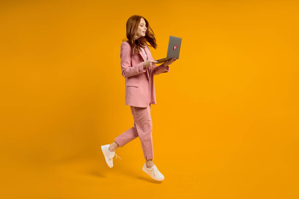 roodharige vrouw run springen typen laptop dragen elegante roze pak geïsoleerd op gele achtergrond - Foto, afbeelding