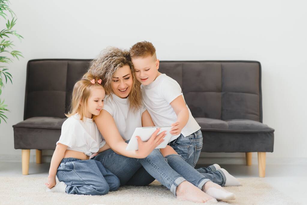 Bela jovem mãe, sua filhinha e filho bonitos estão usando um tablet e sorrindo, sentados no chão em casa - Foto, Imagem