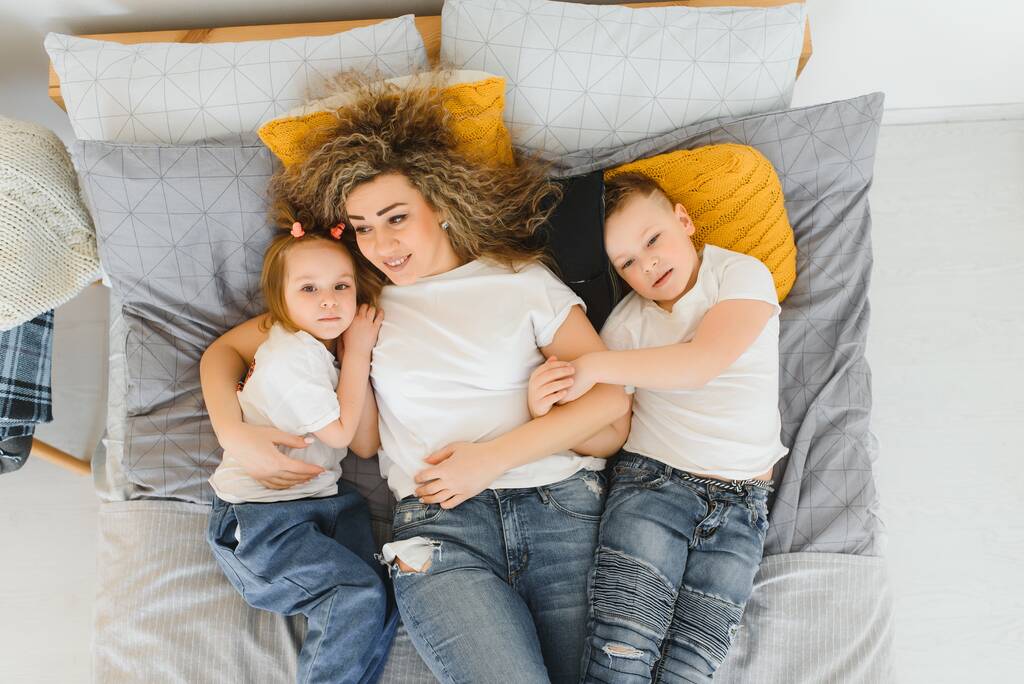 Γελώντας παιδιά παίζουν με τη μητέρα τους ξαπλωμένη σε ένα κρεβάτι στο σπίτι - Φωτογραφία, εικόνα