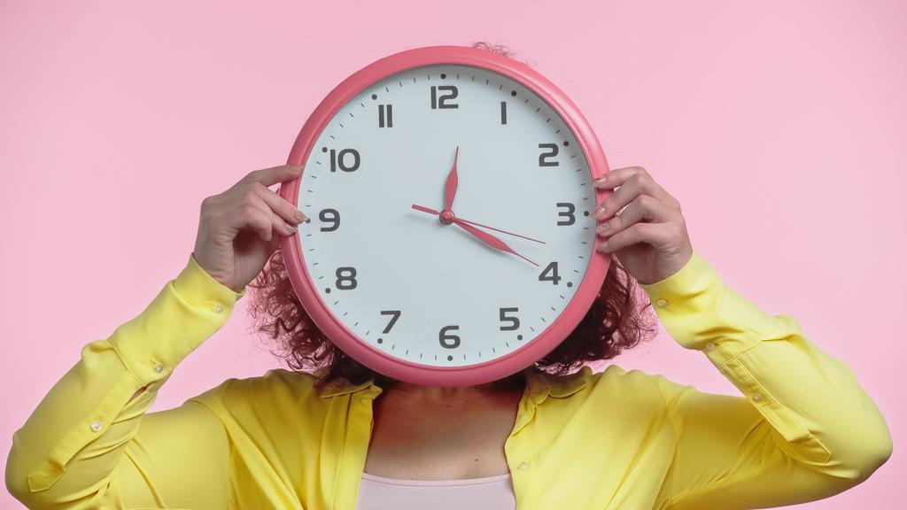 young woman covering face while holding clock isolated on pink  - Photo, Image