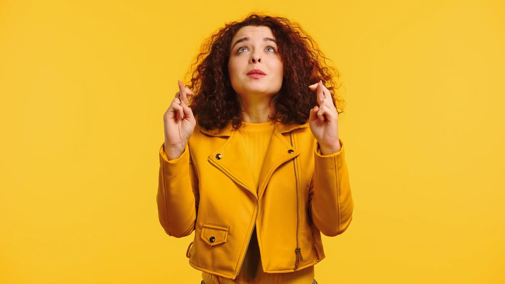молодая кудрявая женщина со скрещенными пальцами загадывает желание и смотрит вверх изолированным на желтый  - Фото, изображение