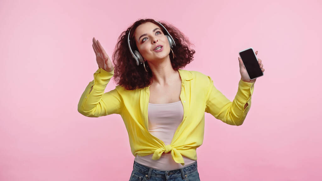 молода жінка в бездротових навушниках тримає смартфон з порожнім екраном ізольовано на рожевому
  - Фото, зображення