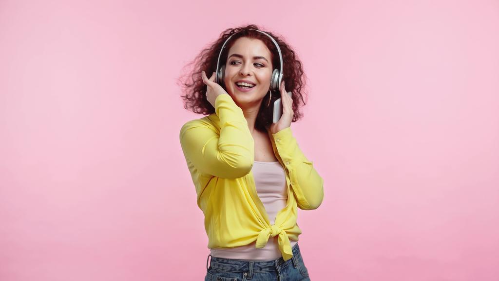 joven mujer alegre en auriculares inalámbricos sosteniendo teléfono inteligente aislado en rosa  - Foto, imagen