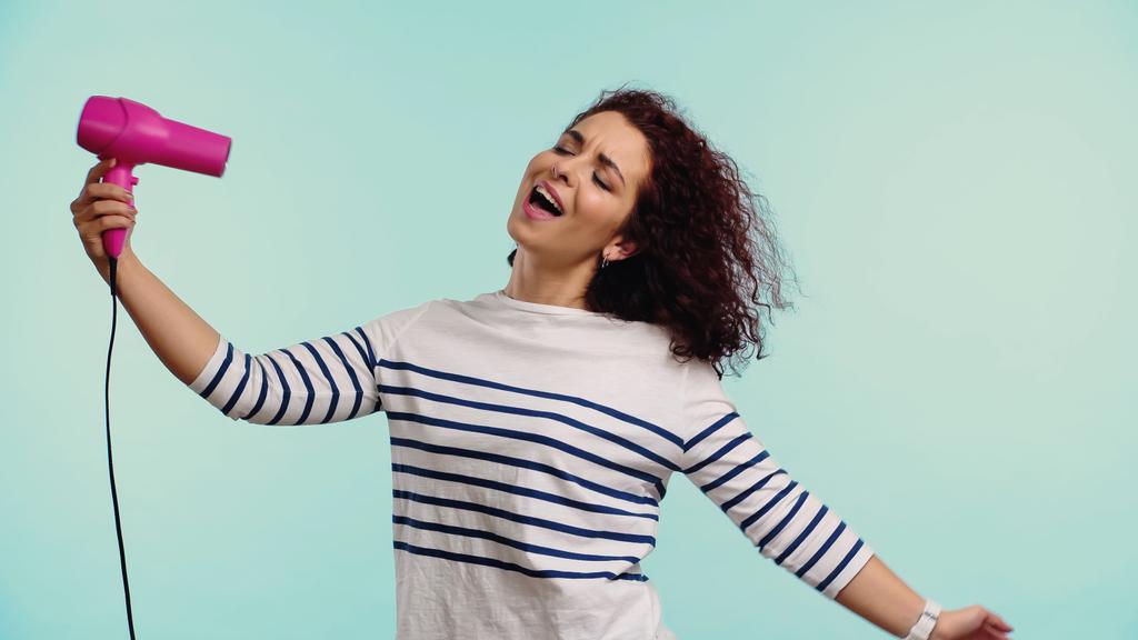 boldog fiatal nő szárítás göndör haj és éneklés elszigetelt kék  - Fotó, kép