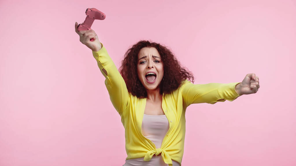 QUIIV, UCRÂNIA - JUNHO 30, 2021: jovem mulher espantada segurando gamepad e alegrando-se isolado em rosa  - Foto, Imagem