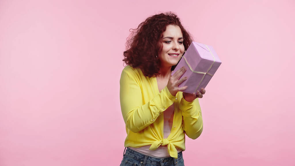 νέα σγουρή γυναίκα κοιτάζοντας τυλιγμένο κουτί δώρου απομονώνονται σε ροζ  - Φωτογραφία, εικόνα