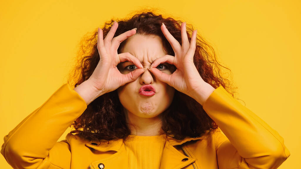 kudrnatá žena napodobující brýle s rukama a šklebící se izolované na žluté - Fotografie, Obrázek