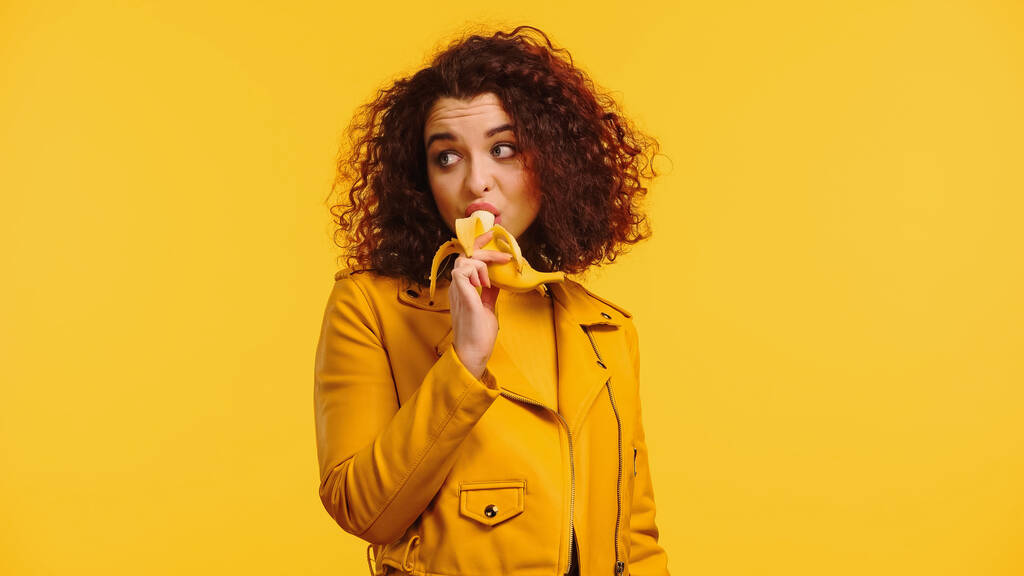 νεαρή γυναίκα με δερμάτινο μπουφάν τρώει μπανάνα απομονωμένη στο κίτρινο  - Φωτογραφία, εικόνα