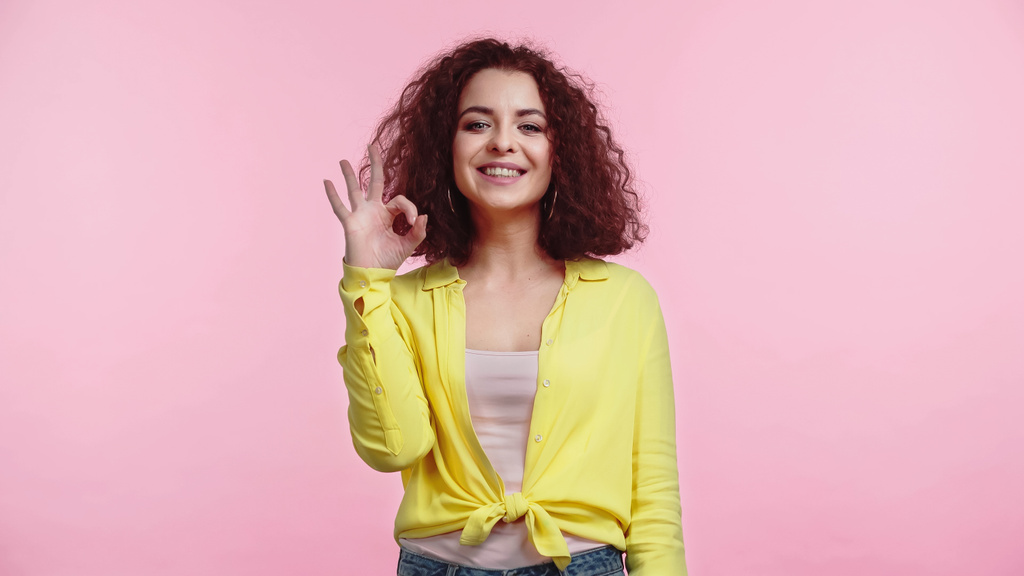 sonriente joven y rizado mujer mostrando ok signo aislado en rosa  - Foto, Imagen