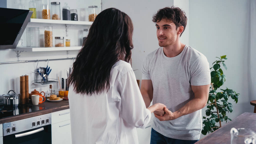 Lächelnder Mann im T-Shirt hält Hand seiner brünetten Freundin in der Küche - Foto, Bild