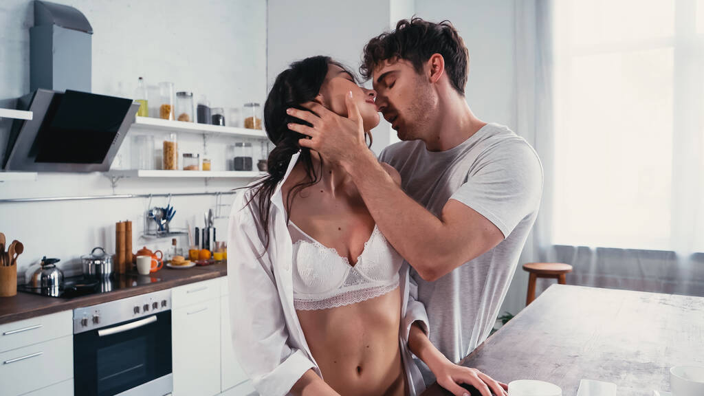 leidenschaftliche Frau in aufgeknöpftem Hemd und BH neben Mann, der sie in der Küche küsst - Foto, Bild