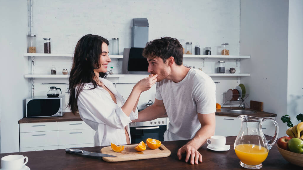 örömteli nő fehér ingben etetés barátja lédús narancs a konyhában - Fotó, kép