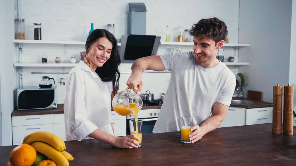 glücklicher Mann gießt frischen Orangensaft neben brünette Frau in weißem Hemd - Foto, Bild