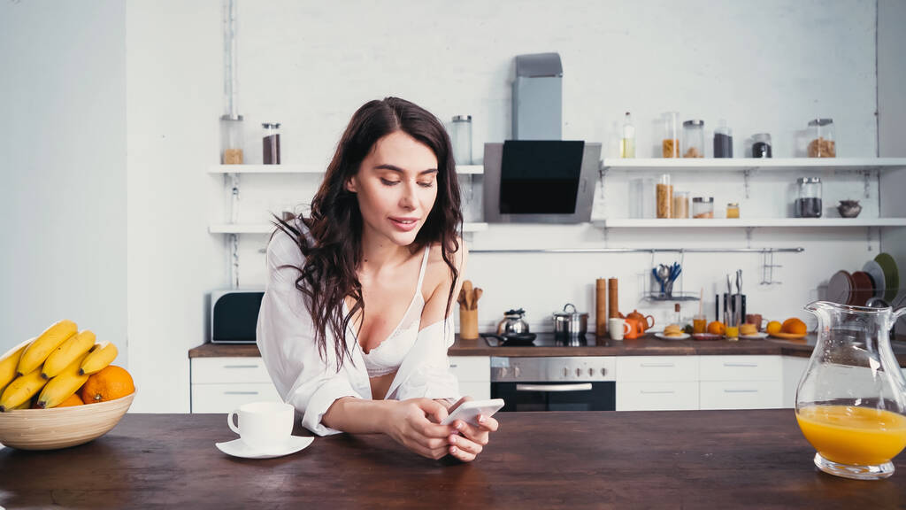 csábító nő fehér ing és melltartó üzenetküldés okostelefon közel kávé és gyümölcs - Fotó, kép