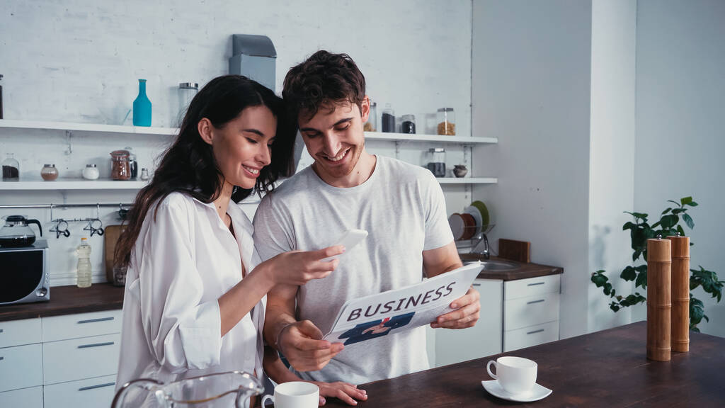 улыбающийся мужчина показывает деловую газету красивой девушке, используя смартфон на кухне - Фото, изображение