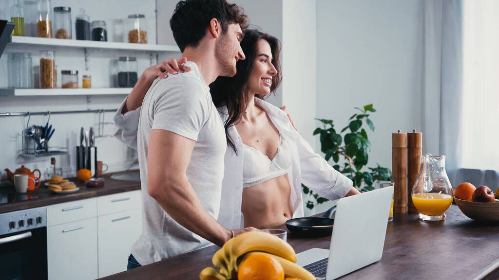 szczęśliwa kobieta w białej koszuli przytula ramię mężczyzny w pobliżu laptopa i owoców w kuchni - Zdjęcie, obraz