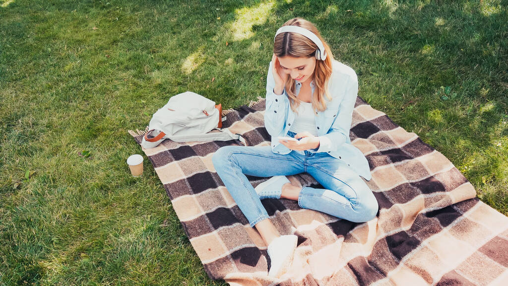 високий кут зору усміхненого студента в бездротових навушниках, використовуючи смартфон на ковдрі в парку
 - Фото, зображення