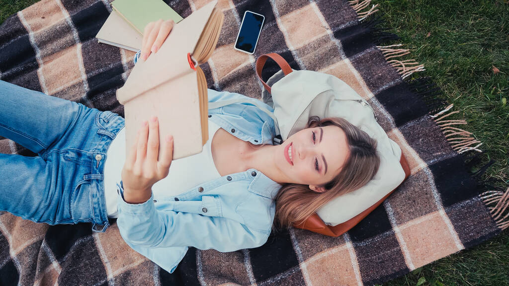 Вгорі вид щасливого студента, який читає книжку і лежить на ковдрі в парку.  - Фото, зображення