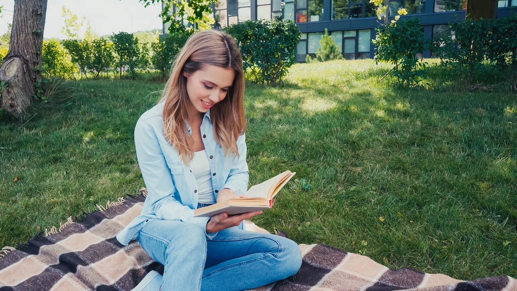 smiling young woman sitting on blanket and reading book in park  - Photo, Image