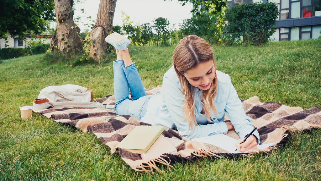 χαρούμενος μαθητής που γράφει στο σημειωματάριο κοντά σε βιβλία, ενώ βρίσκεται σε κουβέρτα στο πάρκο - Φωτογραφία, εικόνα
