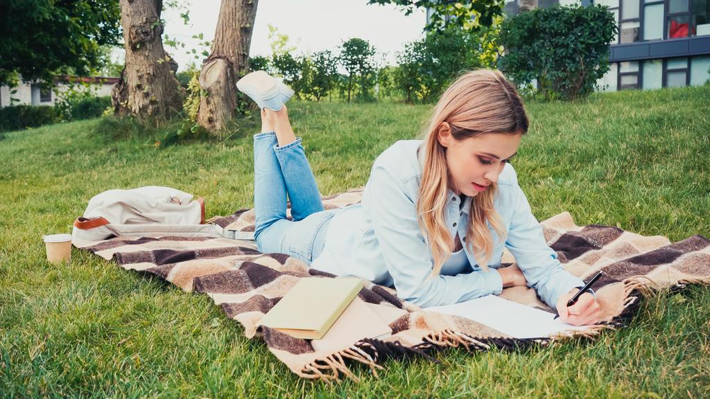étudiant écrit dans un cahier près de livres tout en étant couché sur une couverture dans le parc - Photo, image