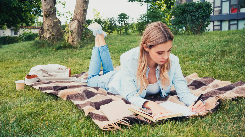 joven estudiante escribiendo en un cuaderno cerca del libro mientras está acostado en una manta en el parque - Foto, Imagen