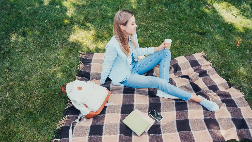 над головой вид счастливого студента, держащего кофе, чтобы подойти к книгам и смартфону на клетчатом одеяле в парке  - Фото, изображение